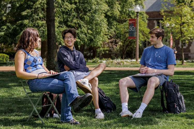 three students sitting in chairs outside on campus